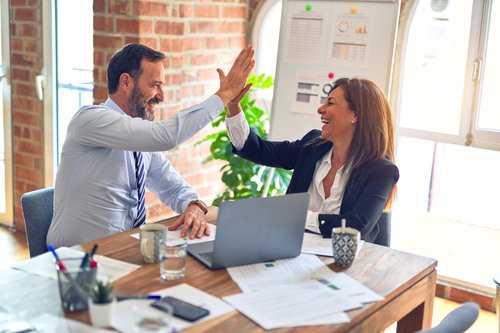 Man and woman in an office at a table with a laptop, high-fiving