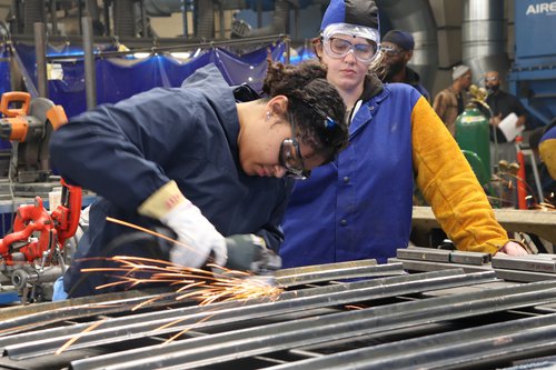 Two people wearing goggles; one is using a power tool