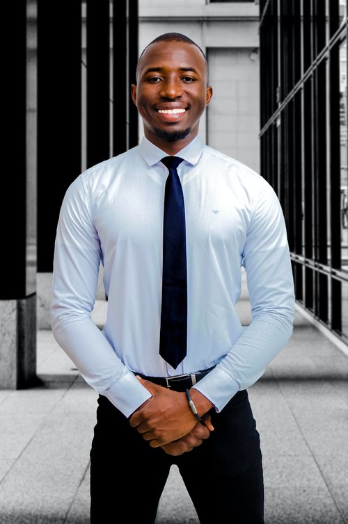 Black man smiling, standing with hands clasped, wearing a collared shirt and tir.